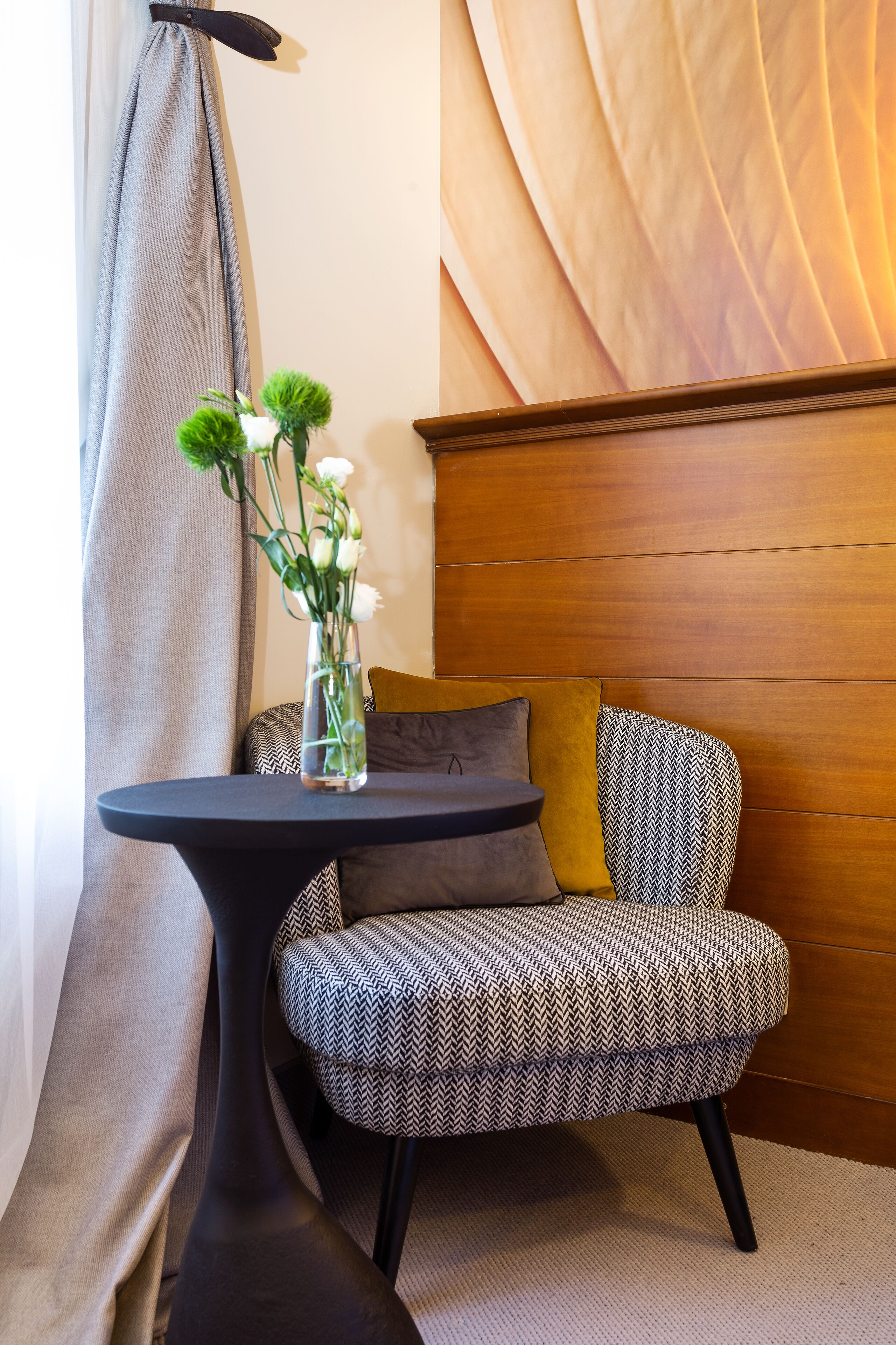 Una habitación de esquina con una mesa redonda, una silla estampada, un florero con flores, una lámpara y un cabecero de madera.