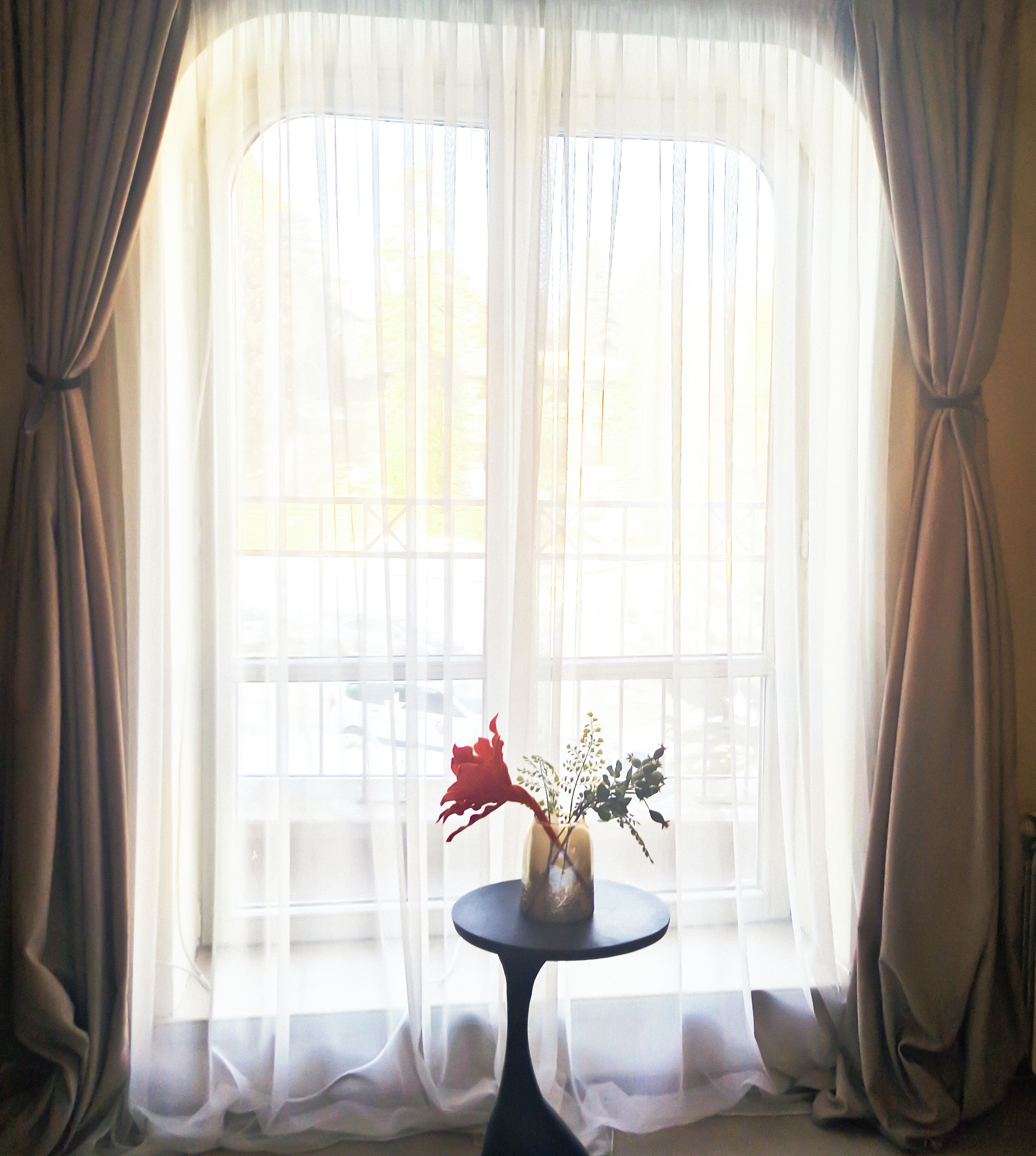 A room with a large window covered by sheer curtains, a small round table with a vase of flowers, and a view of the outside.