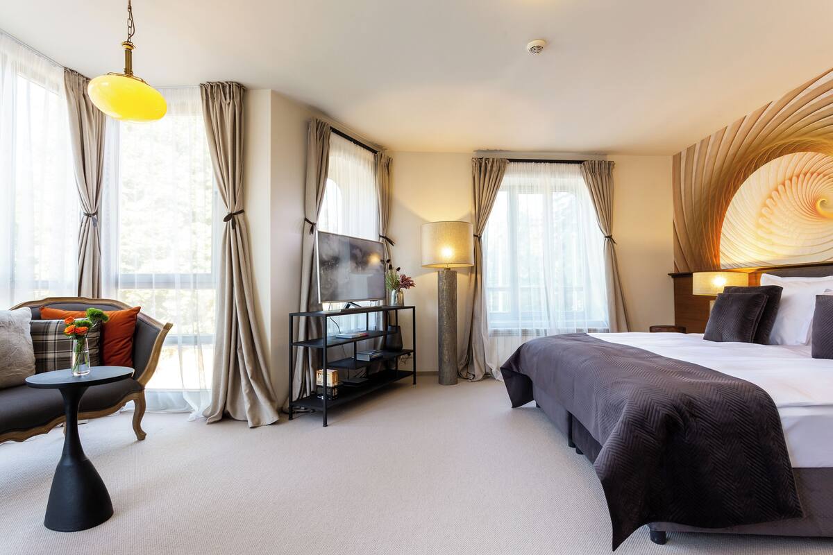 A hotel room with a bed, a sofa, a TV on a stand, a lamp, and curtains.