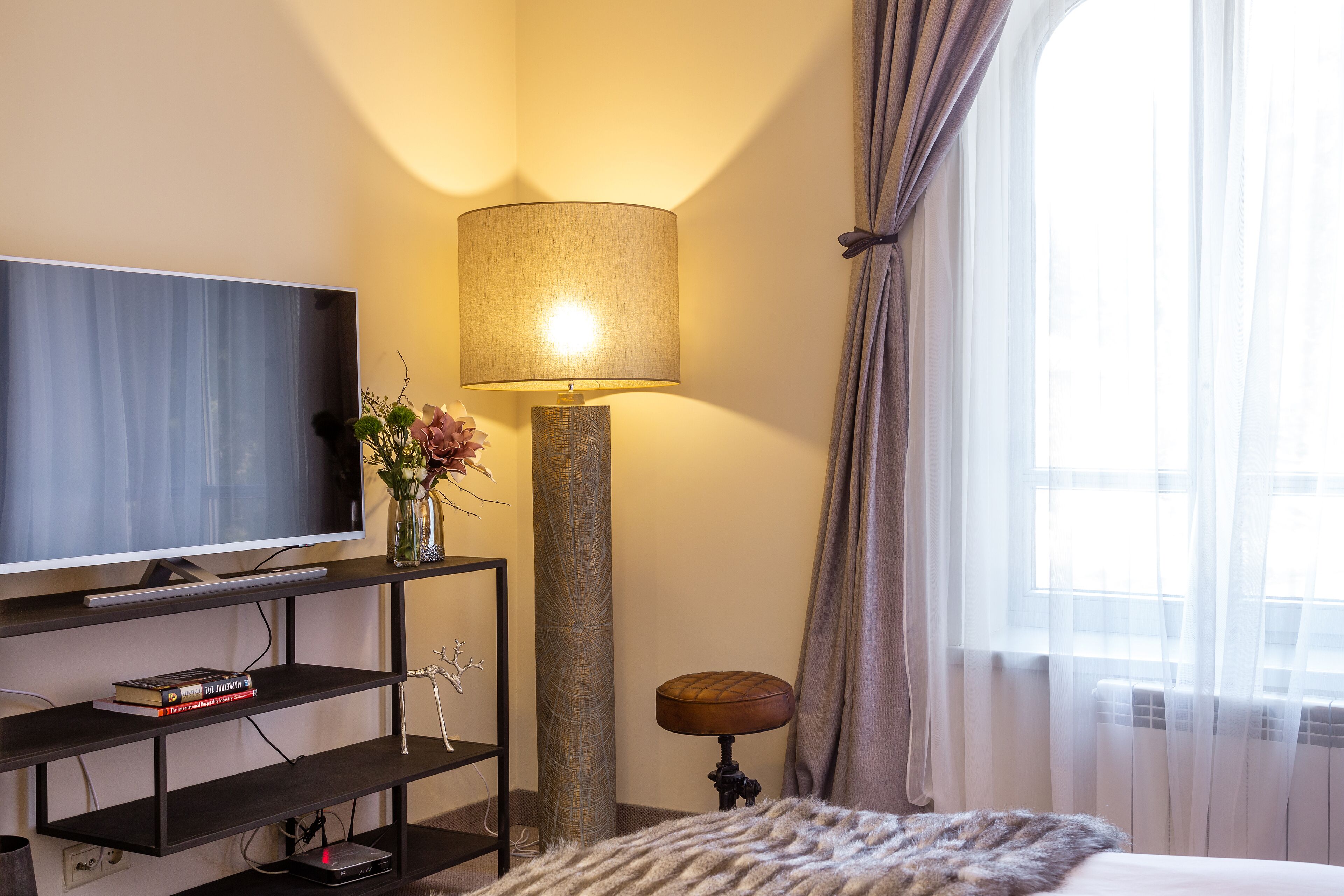 Hotelkamer met een flatscreen televisie op een donkere houten standaard, een hoge vloerlamp, een klein rond krukje en een groot raam met dunne gordijnen.