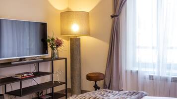 Hotelkamer met een flatscreen televisie op een donkere houten standaard, een hoge vloerlamp, een klein rond krukje en een groot raam met dunne gordijnen.