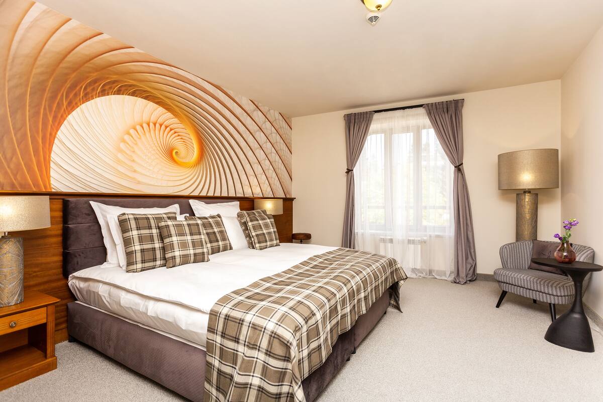 A hotel room with a large bed, a headboard with a unique design, a side table with a lamp, a chair, and a window with curtains.