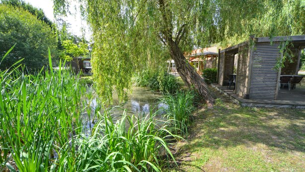 A serene garden with a pond, lush greenery, and a wooden structure with outdoor seating.