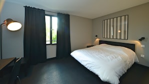 A hotel room with a bed, a desk, a chair, a window with curtains, and a wall-mounted lamp.