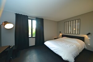 Una habitación de hotel con cama, escritorio, silla, ventana con cortinas y una lámpara fijada a la pared.