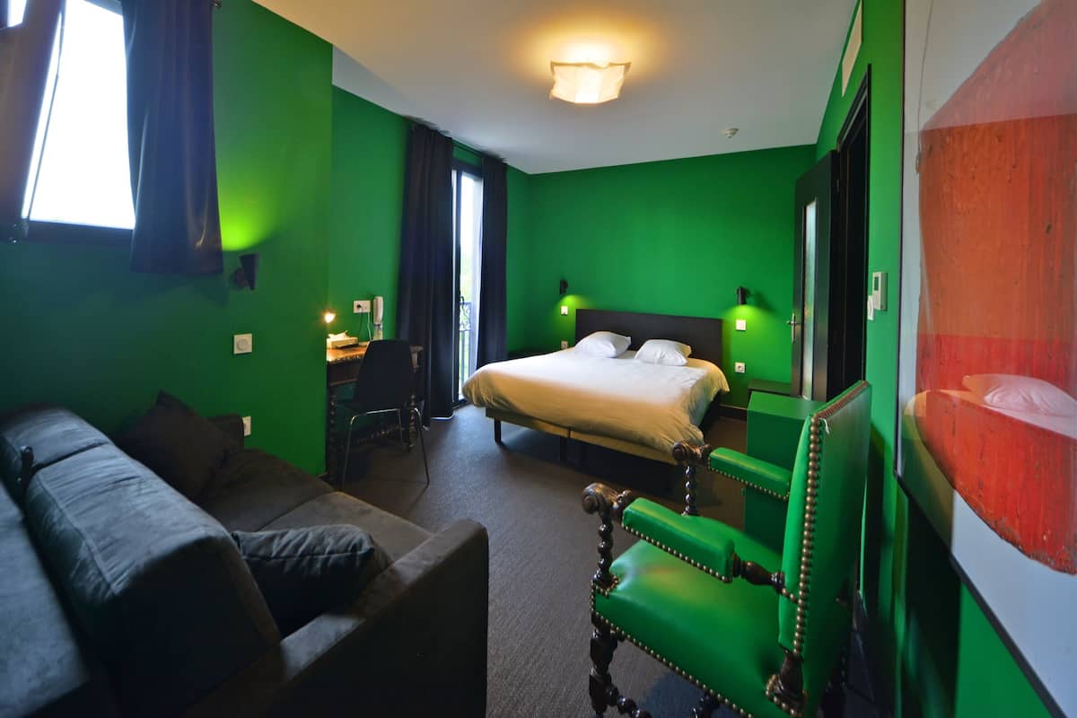 A hotel room with a green wall, a bed, a green chair, a desk, and a window with curtains.