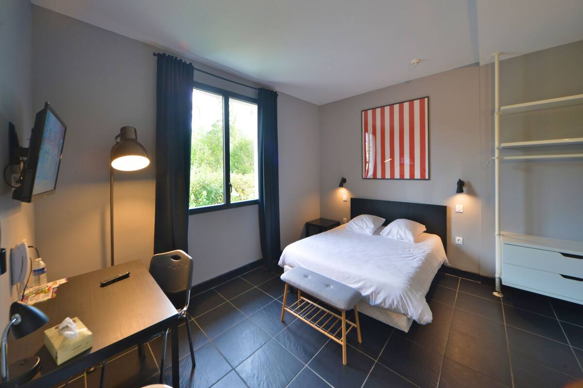 A hotel room with a bed, bedside tables, a desk with a lamp, a chair, a window with curtains, and a wall-mounted shelf.