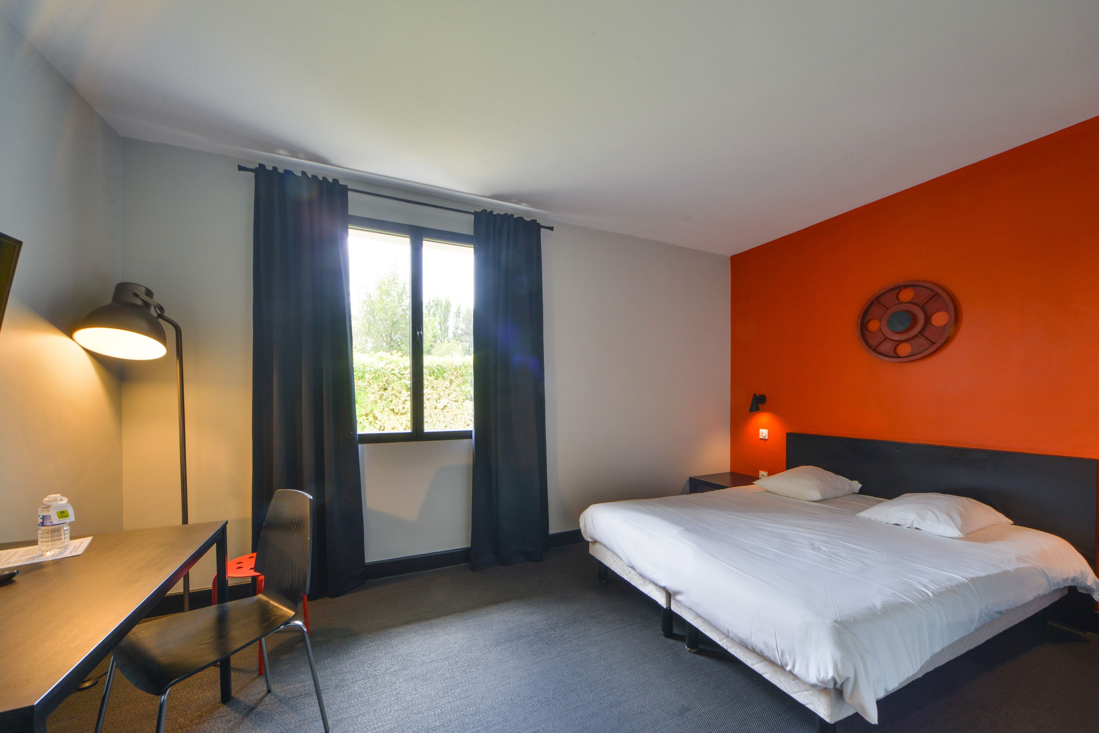 Quarto de hotel com cama, escrivaninha, cadeira, abajur e janela com cortinas.
