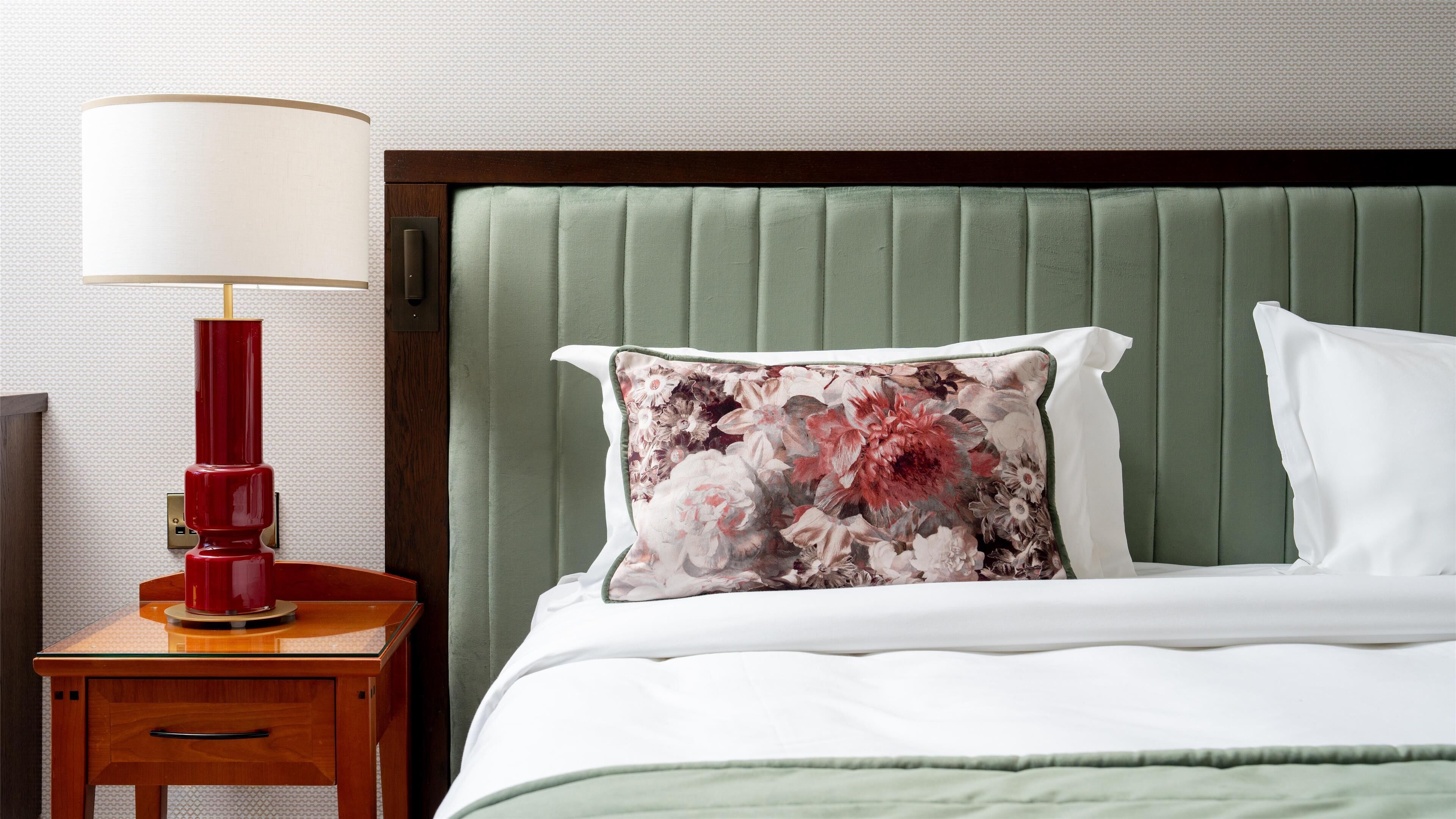 Un lit bien fait, avec une tête de lit verte, un coussin à motifs floraux et un coussin blanc. Une table de chevet en bois avec une lampe rouge se trouve à côté du lit.