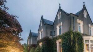 A large, stately building with a formal entrance, manicured hedges, and a paved pathway leading to a seating area.