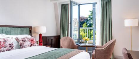 A hotel room with a bed, two chairs, a small table with a vase, and a view of a balcony with greenery.