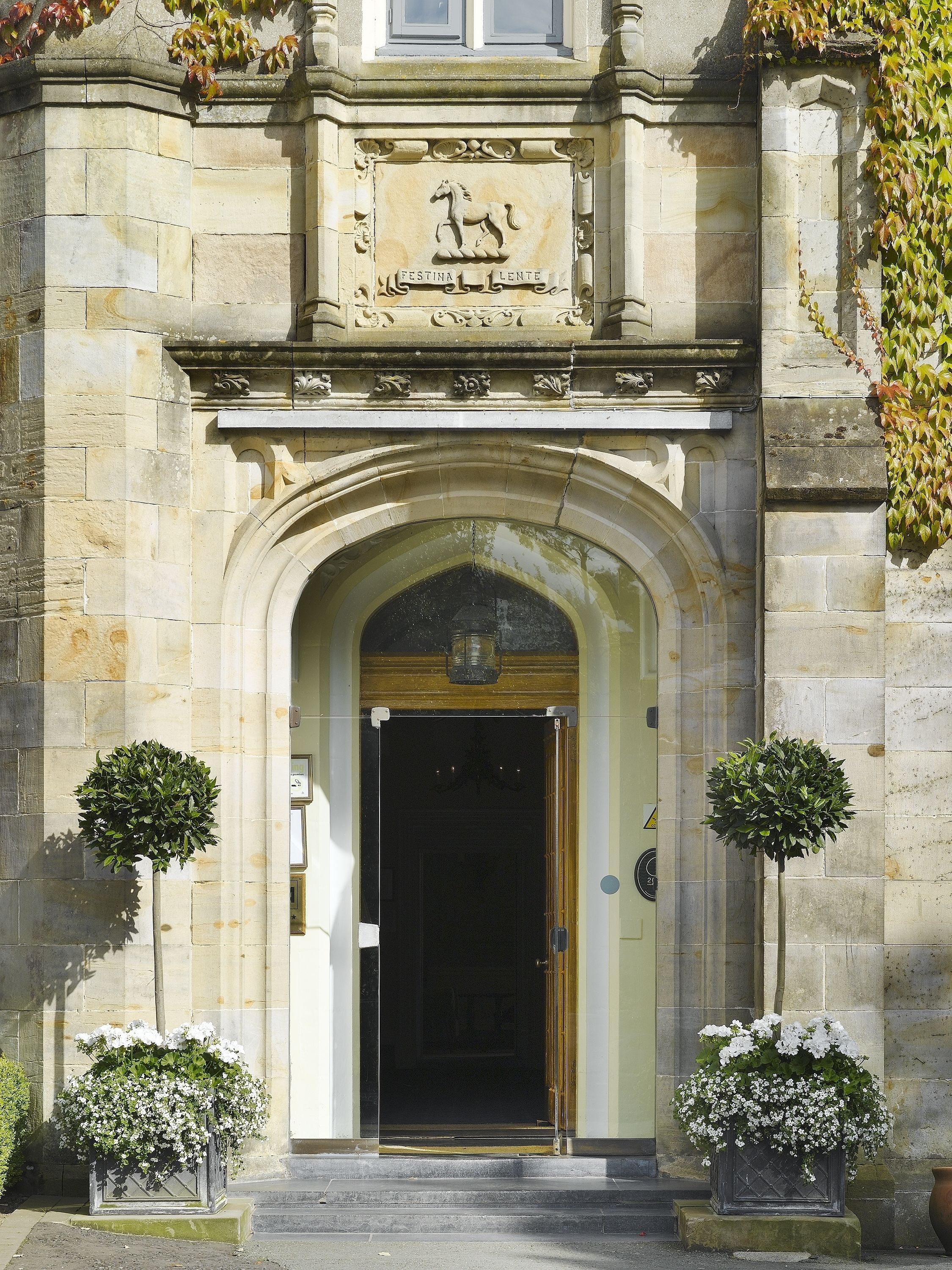 A stone building entrance with a coat of arms, an arched doorway, and two potted plants.