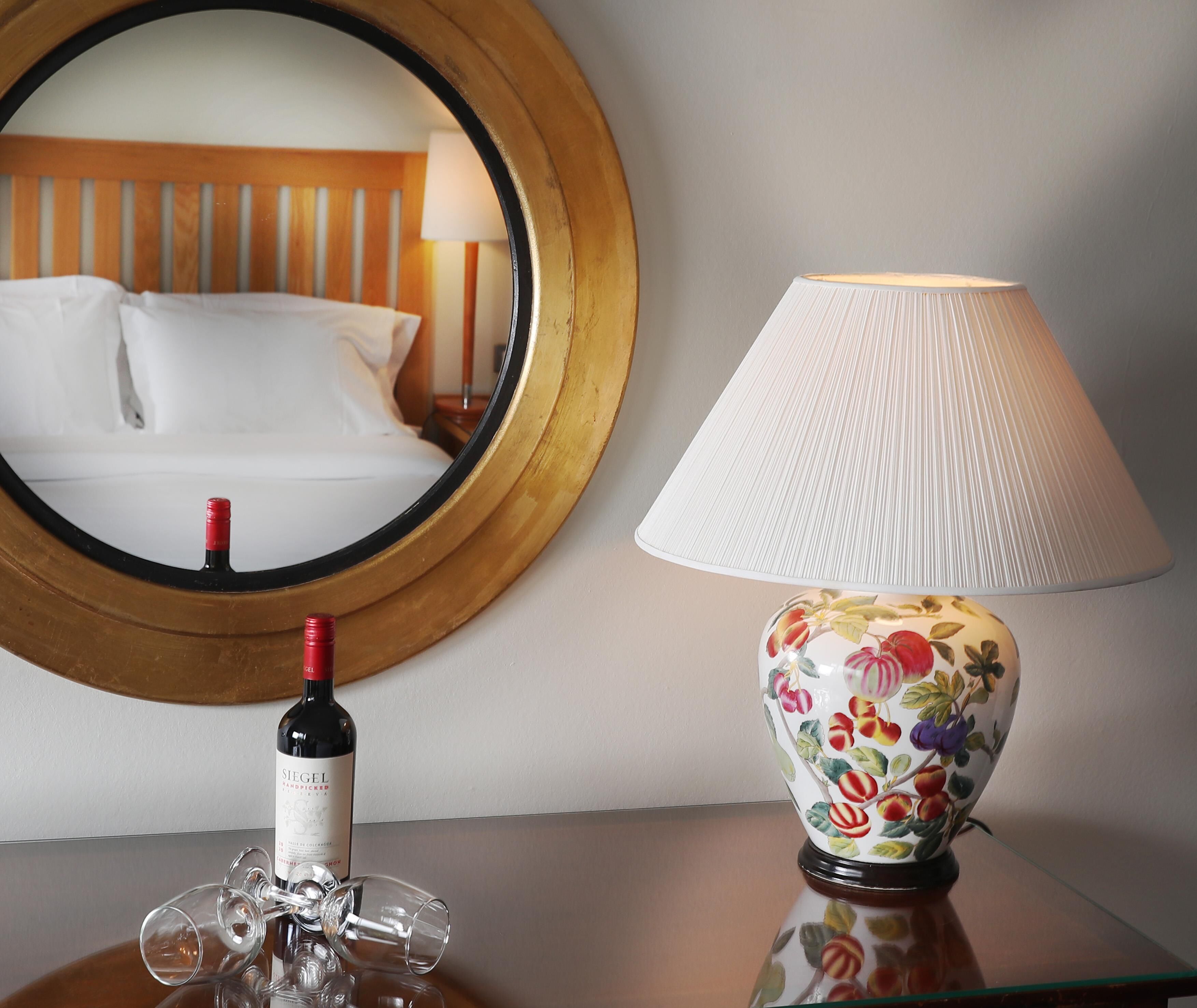 Habitación de hotel con una cama, un espejo redondo, una mesa con una botella de vino y copas, y una lámpara con motivos florales.