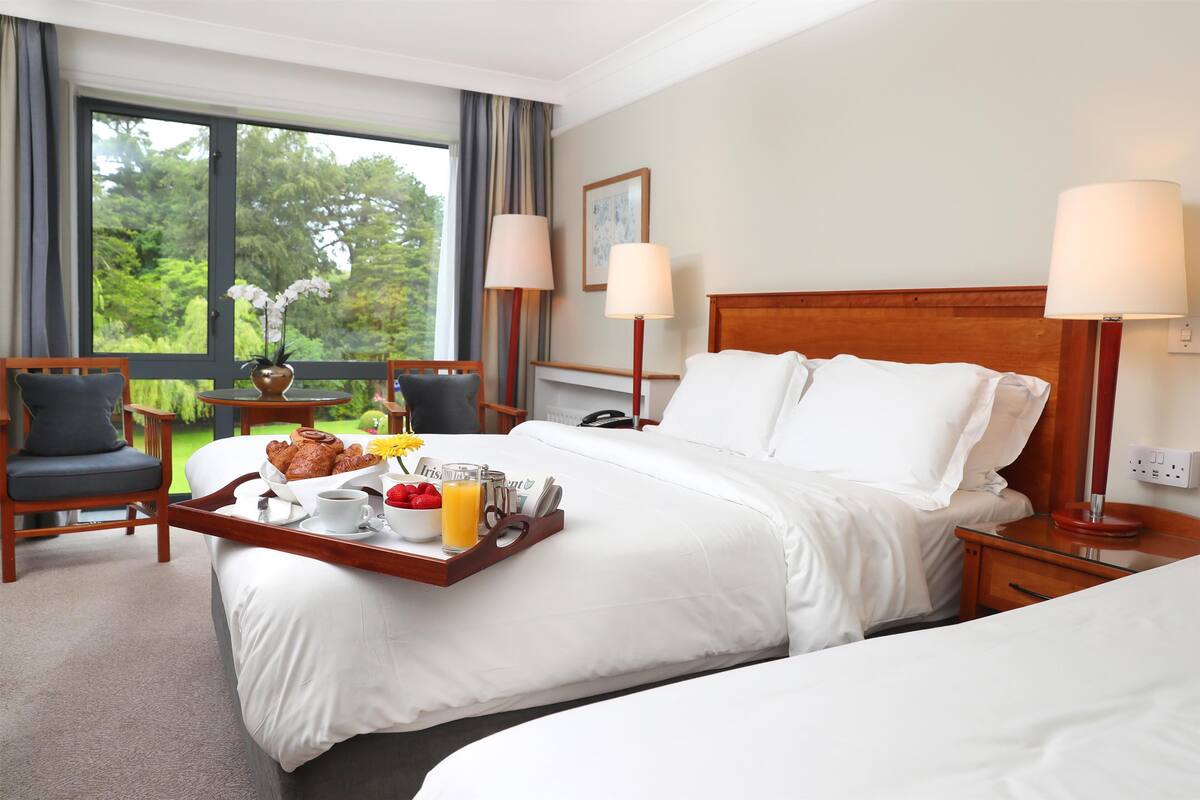 Une chambre d’hôtel avec un grand lit, deux tables de chevet, une chaise, une fenêtre avec des rideaux et un plateau contenant des articles pour le petit-déjeuner.