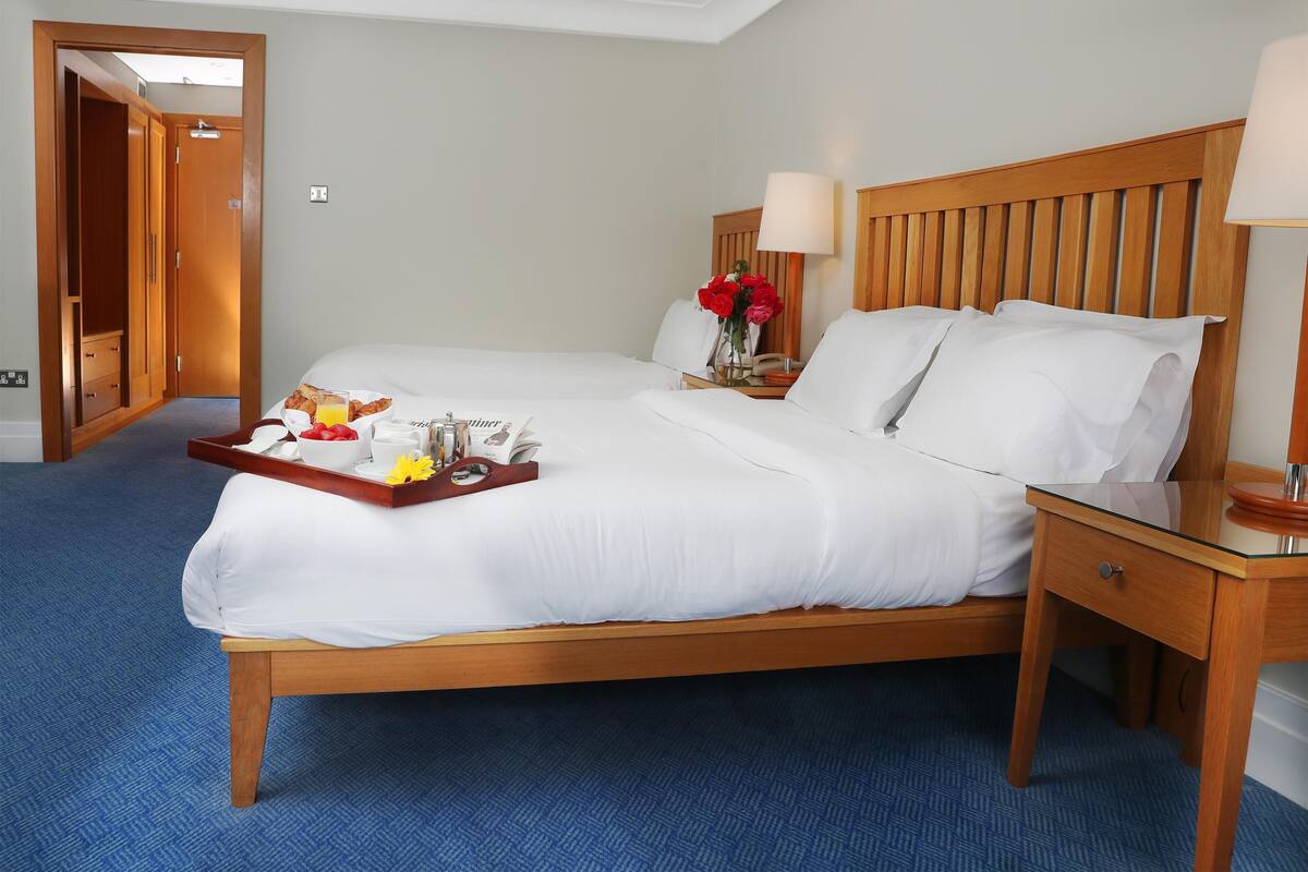 Une chambre d’hôtel avec un lit en bois, une table de chevet avec une lampe, un plateau contenant des articles pour le petit-déjeuner et une porte donnant sur une autre pièce.