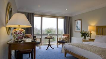 Habitación de hotel con una cama grande, un escritorio con lámpara, una silla y vistas al exterior a través de amplios ventanales.
