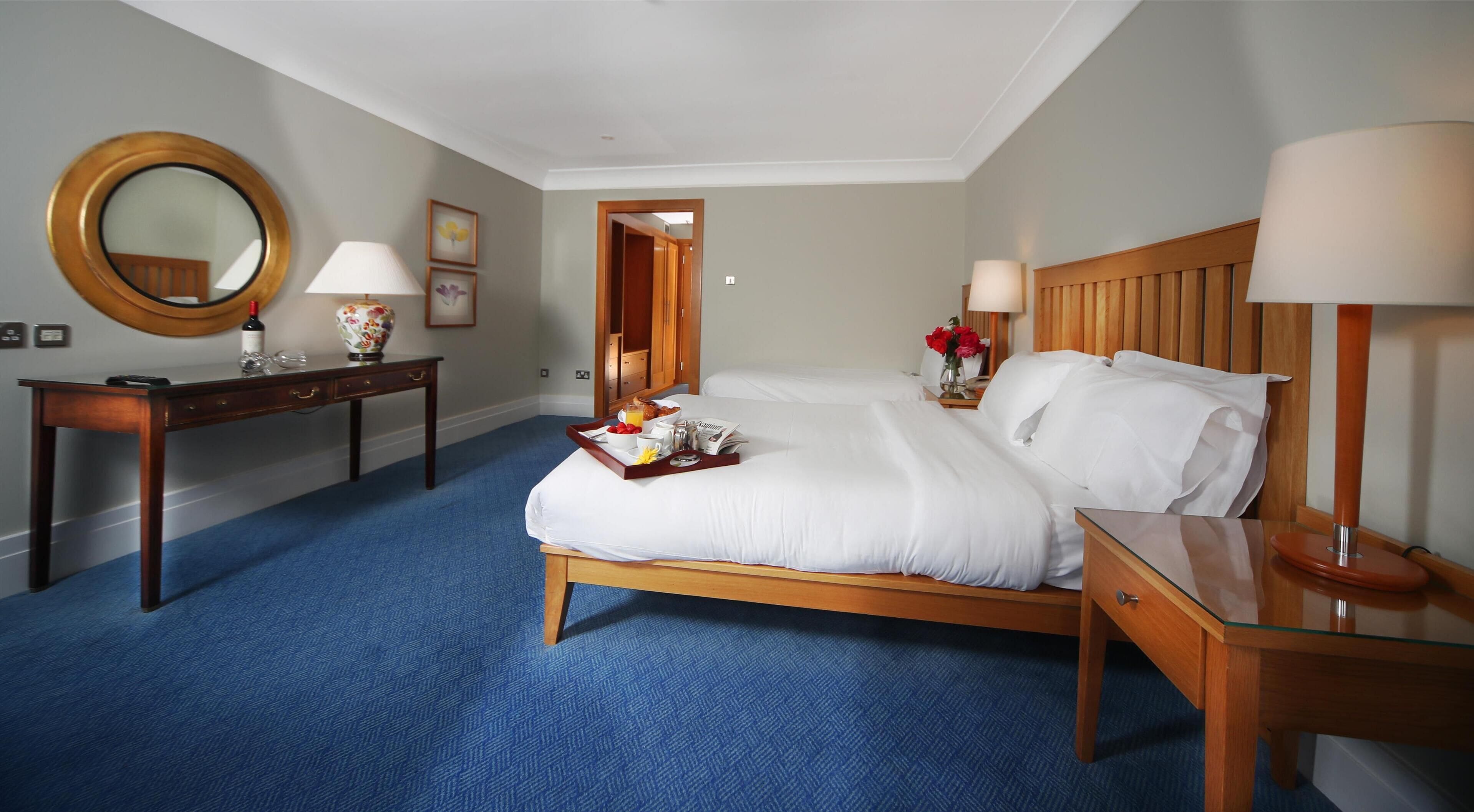 Habitación de hotel con una cama grande, una mesita de noche de madera, un escritorio con espejo y una alfombra azul.