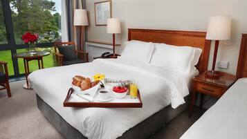 Habitación de hotel con una cama grande, una mesita de noche con lámpara, una pequeña mesa con un jarrón de flores y una bandeja de desayuno sobre la cama.