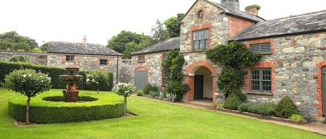 A stone building with a garden in front, including a fountain and trimmed hedges.