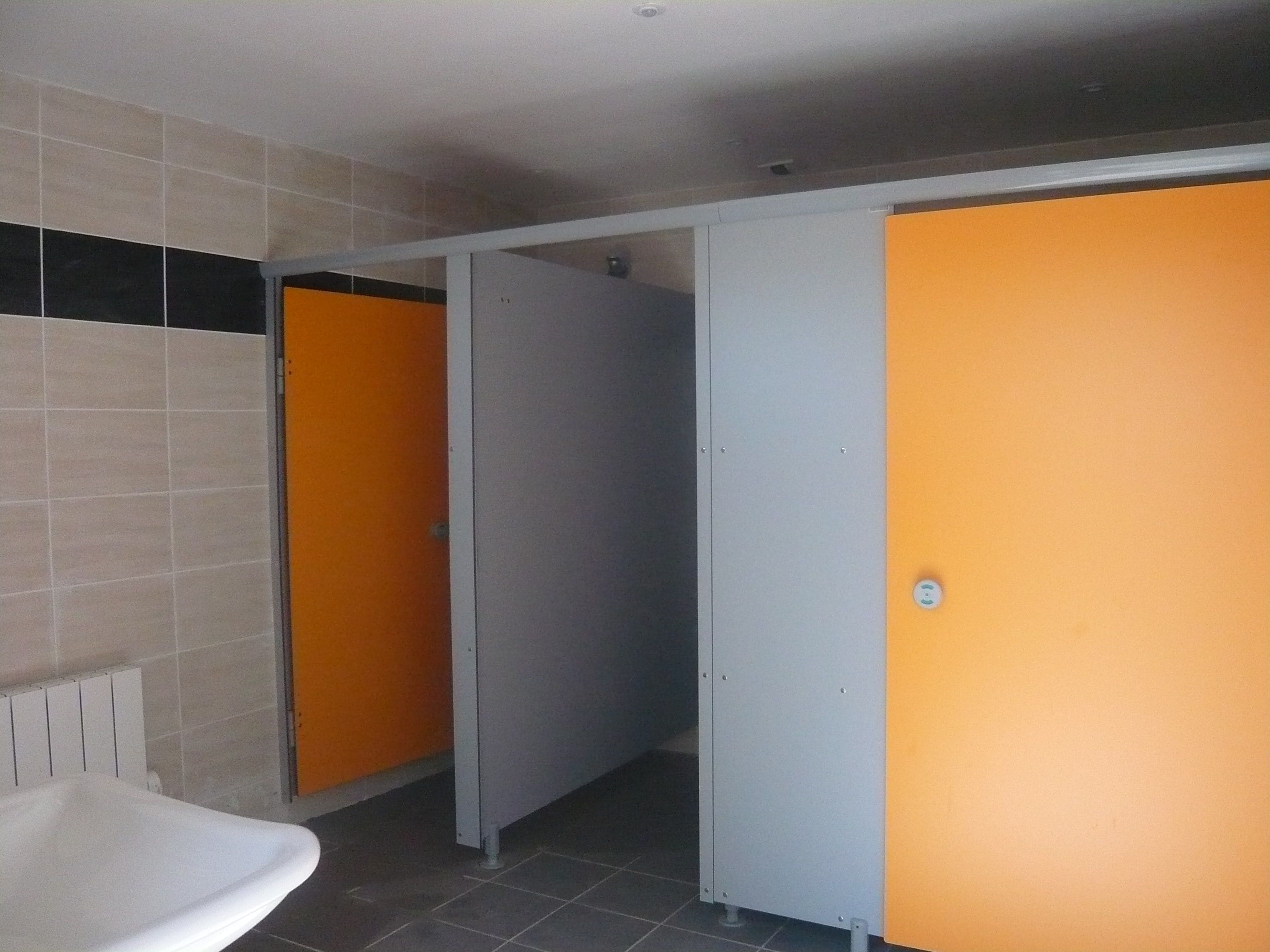 Un baño con un lavamanos blanco, paredes de azulejos y tres puertas, dos de color naranja y una gris.