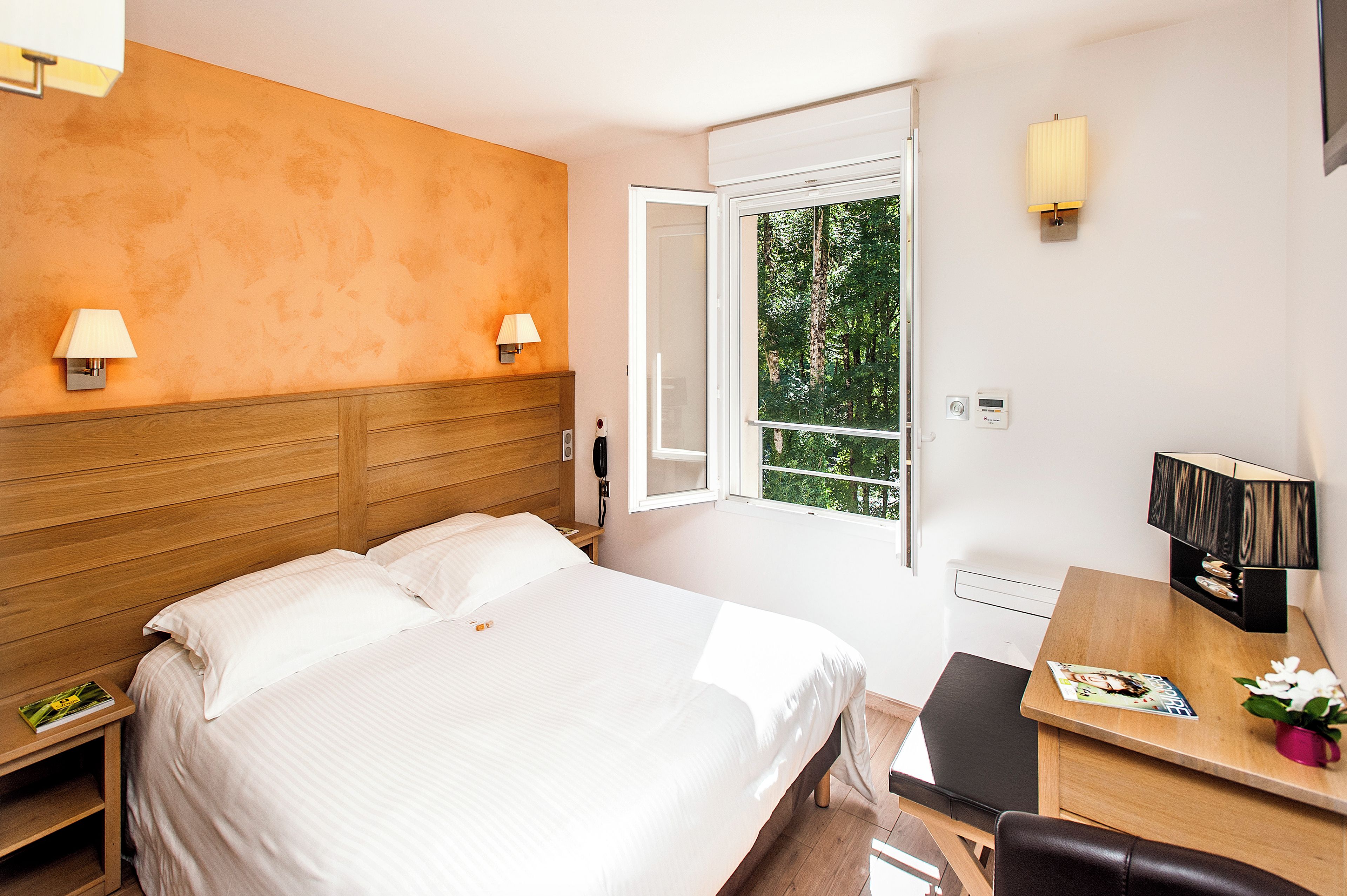 Hotelkamer met een bed, nachtlampjes, een bureau met stoel, een raam met uitzicht op bomen en een tafeltje met een tijdschrift en een bloem.