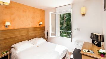Hotelkamer met een bed, nachtlampjes, een bureau met stoel, een raam met uitzicht op bomen en een tafeltje met een tijdschrift en een bloem.