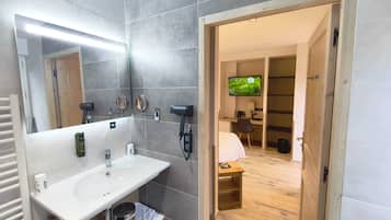 Ein modernes Badezimmer mit einem großen Spiegel, einem Waschbecken und einem Haartrockner. Im angrenzenden Raum befindet sich ein Fernseher.