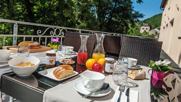 Una mesa para comer al aire libre con alimentos para el desayuno, como pan, fruta y bebidas, con árboles y un edificio de fondo.