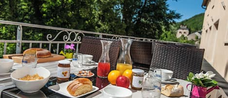 An outdoor dining table with breakfast items, including bread, fruit, and beverages, set against a backdrop of trees and a building.