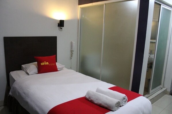 A hotel room with a bed, a red pillow, a white bedspread, and a red and white towel.