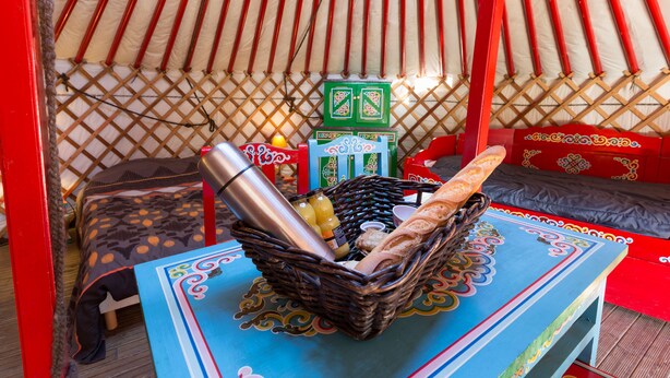 Interno di una yurta tradizionale, con un cesto intrecciato contenente pane, una termica e una bottiglia su un tavolo blu.