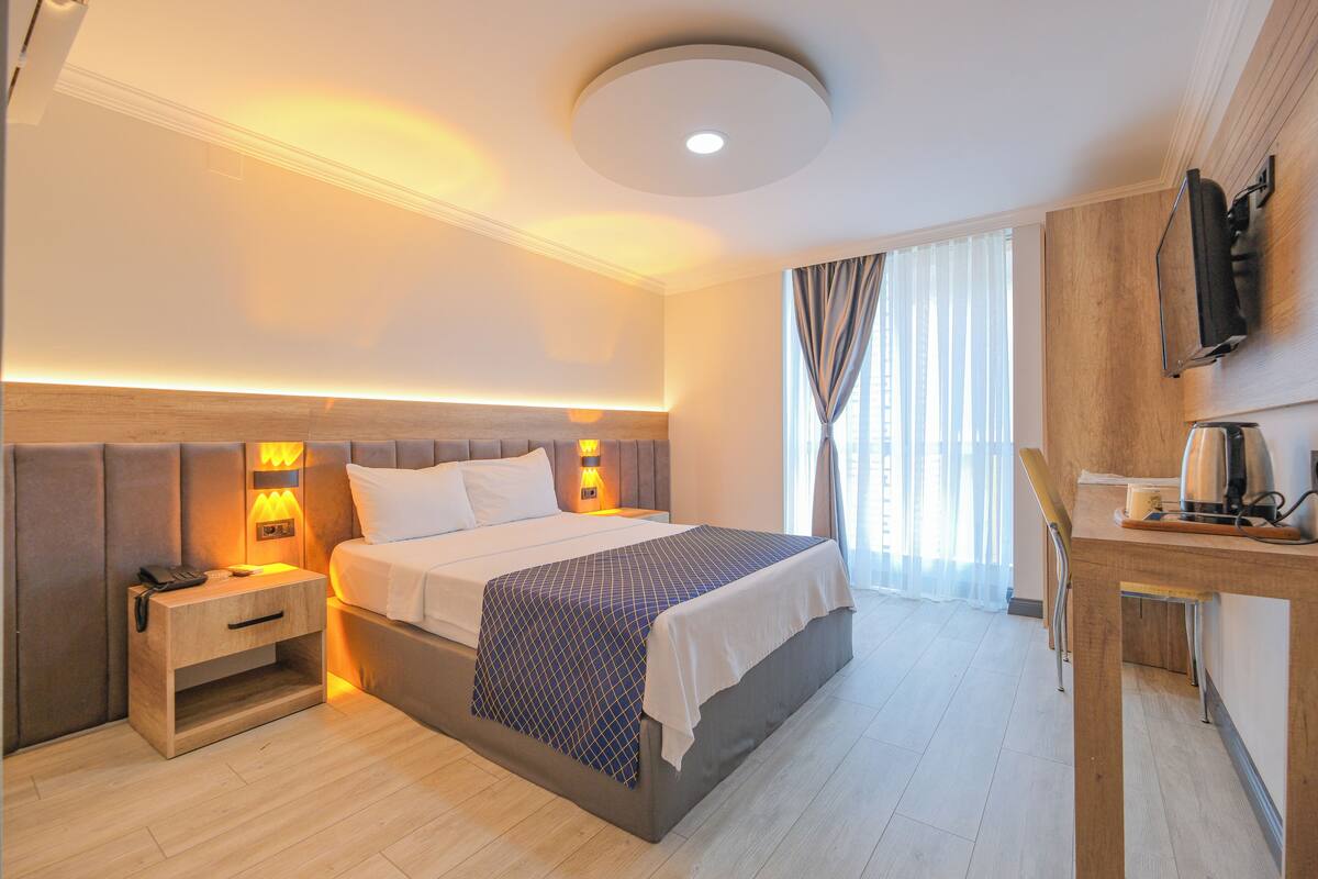 Una habitación de hotel moderna con una cama grande, lámparas de noche, una mesita de noche de madera, un escritorio con una silla y un televisor de pantalla plana montado en la pared.