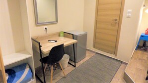 A small, compact room with a wooden desk, a chair, a compact fridge, a trash bin, a mirror, and a wooden door. - Hotel Achillion (Piraeus)
