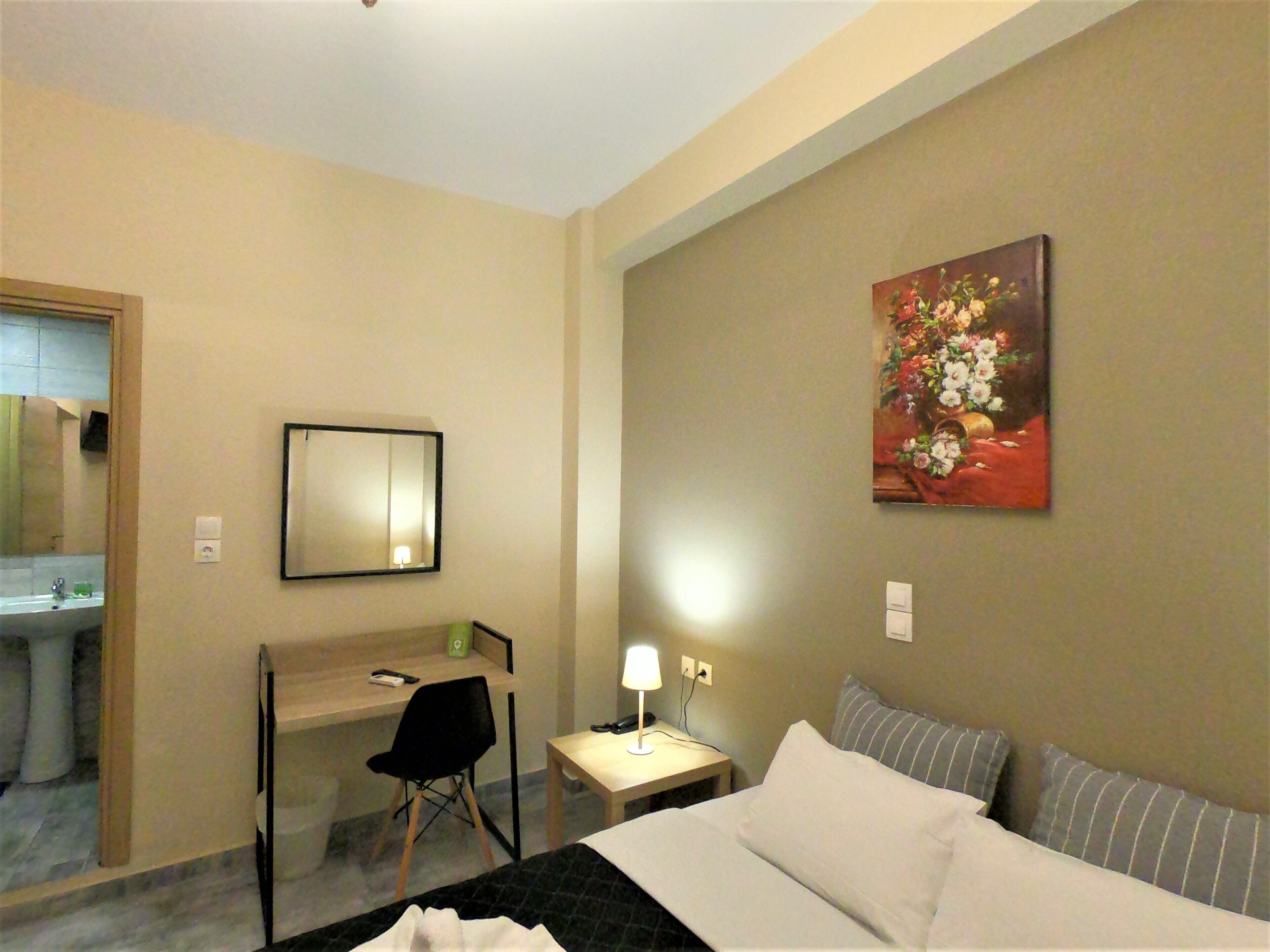 Habitación de hotel con cama, escritorio con silla, pequeña mesa con lámpara, un cuadro en la pared y baño visible a través de una puerta abierta.