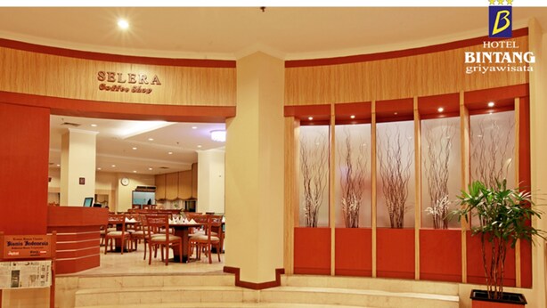 Um saguão de hotel com uma mesa de recepção, uma cafeteria chamada 'Selera Coffee Shop' e uma escadaria que leva ao andar superior.
