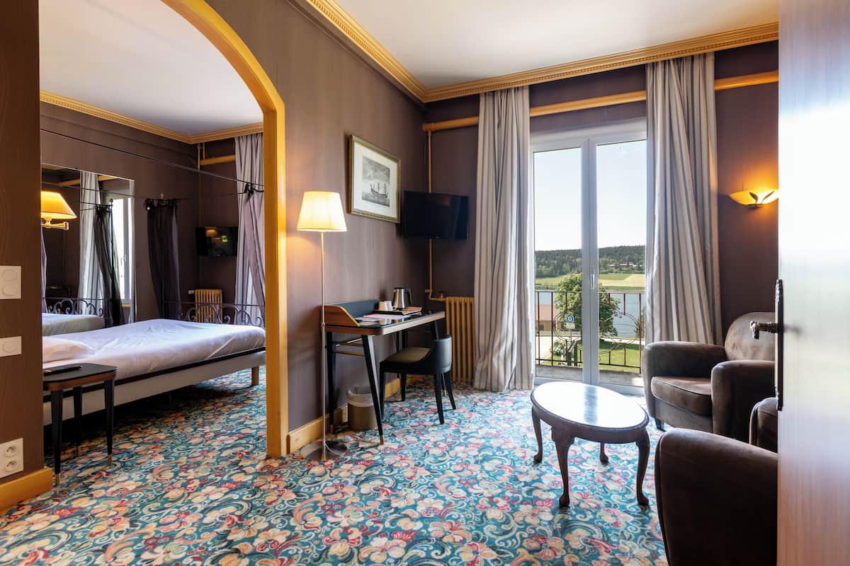 A hotel room with a bed, desk, chair, and a small table. There is a TV mounted on the wall and a window with curtains.