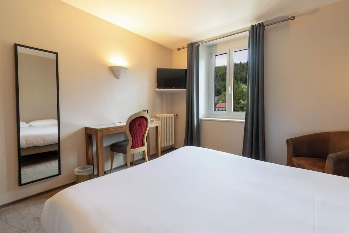 A hotel room with a large bed, a desk with a chair, a television mounted on the wall, and a window with curtains.