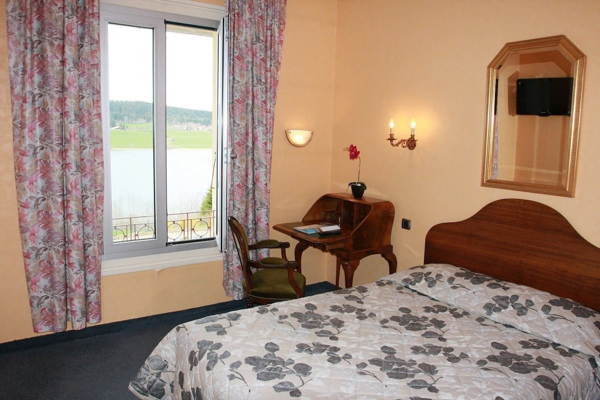 A bedroom with a bed, a desk with a chair, a window with curtains, a wall-mounted lamp, and a mirror.