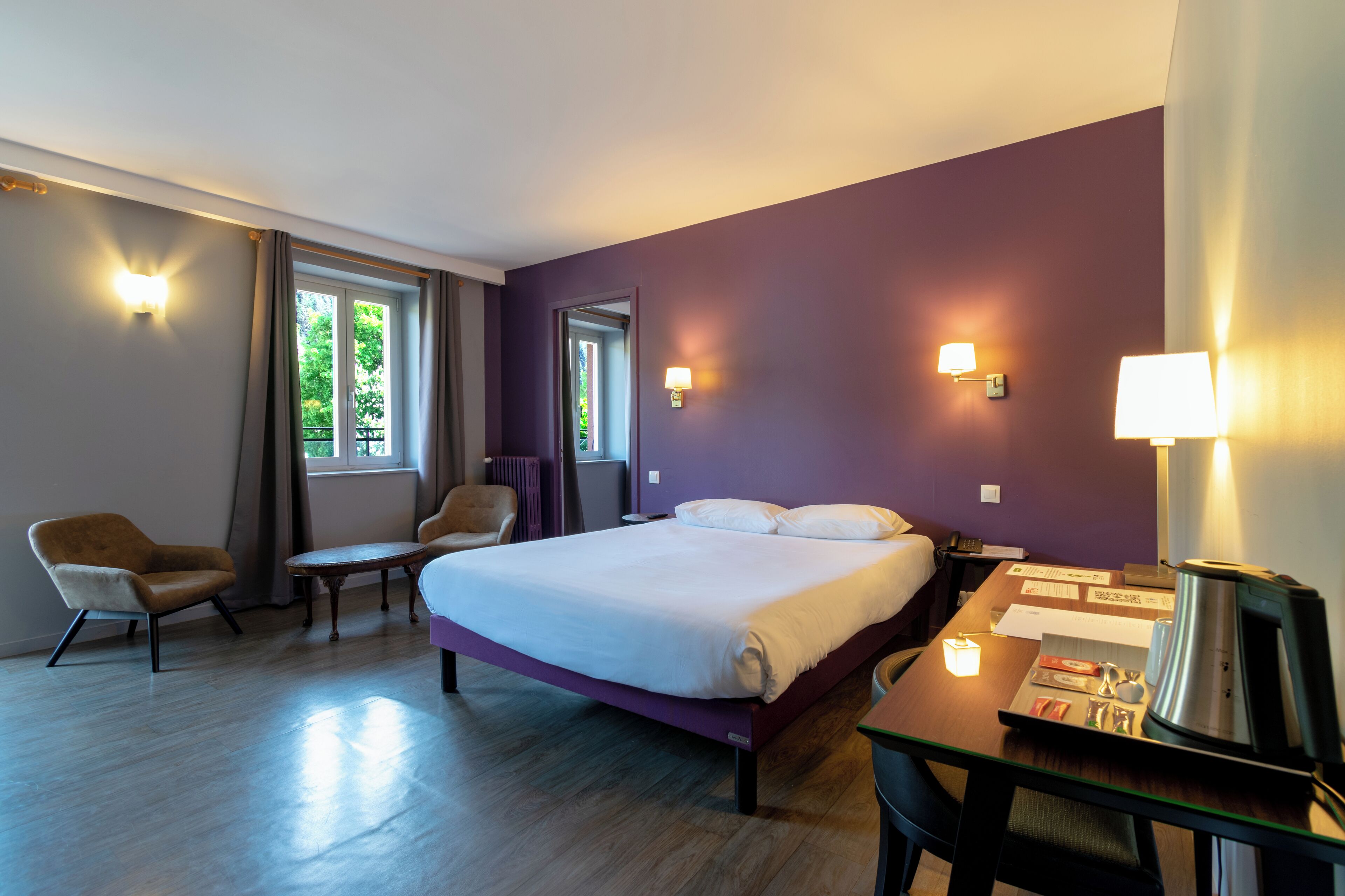 Habitación de hotel con una cama grande, un escritorio con cafetera, una silla y dos lámparas de pared.