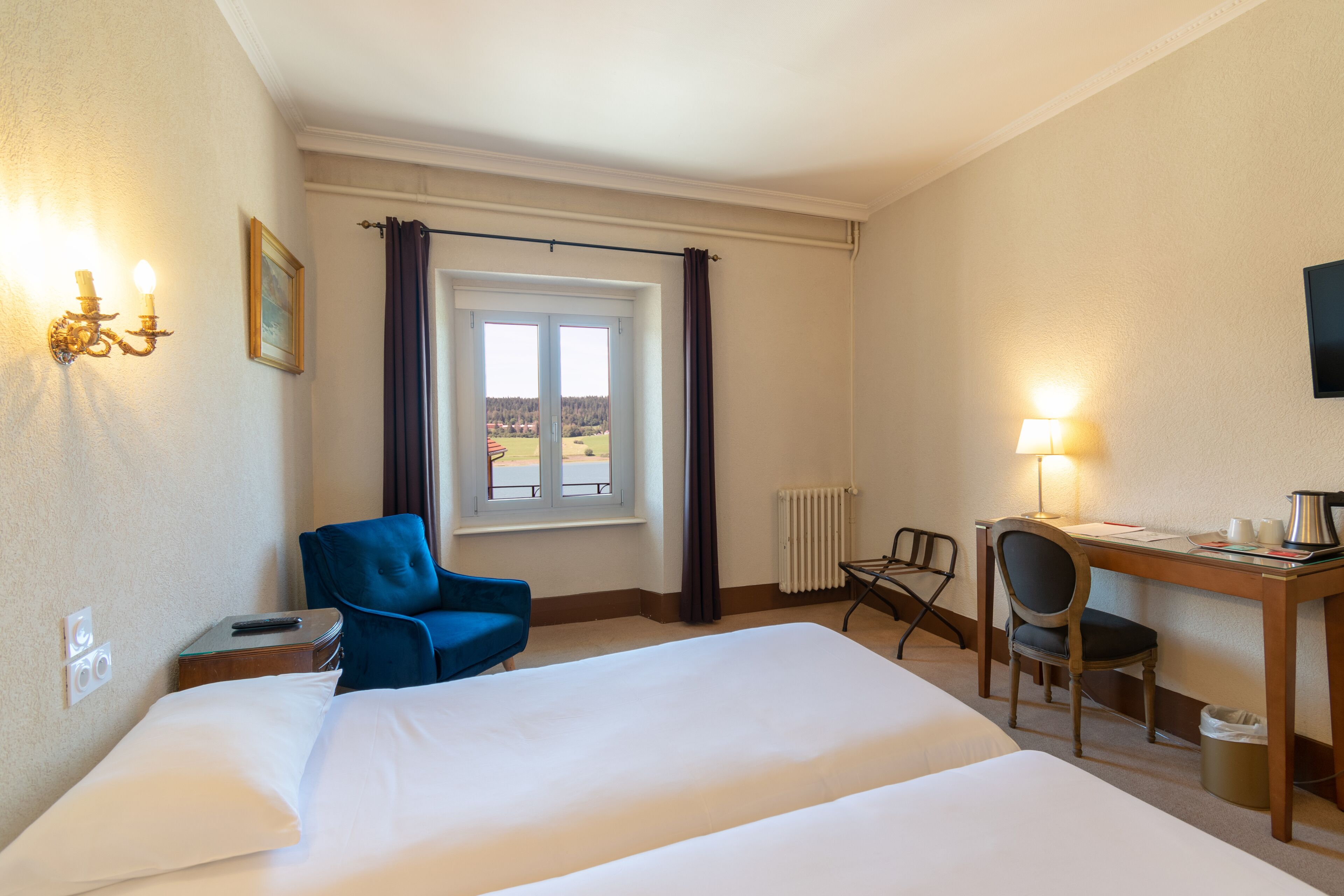 Habitación de hotel con una cama grande, un escritorio con una silla, un sillón azul, una ventana con cortinas y una lámpara de pared.