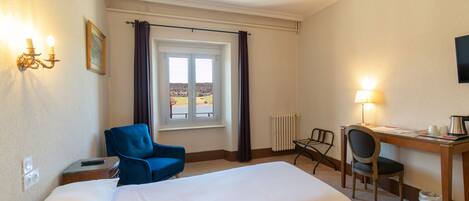 Habitación de hotel con una cama grande, un escritorio con una silla, un sillón azul, una ventana con cortinas y una lámpara de pared.