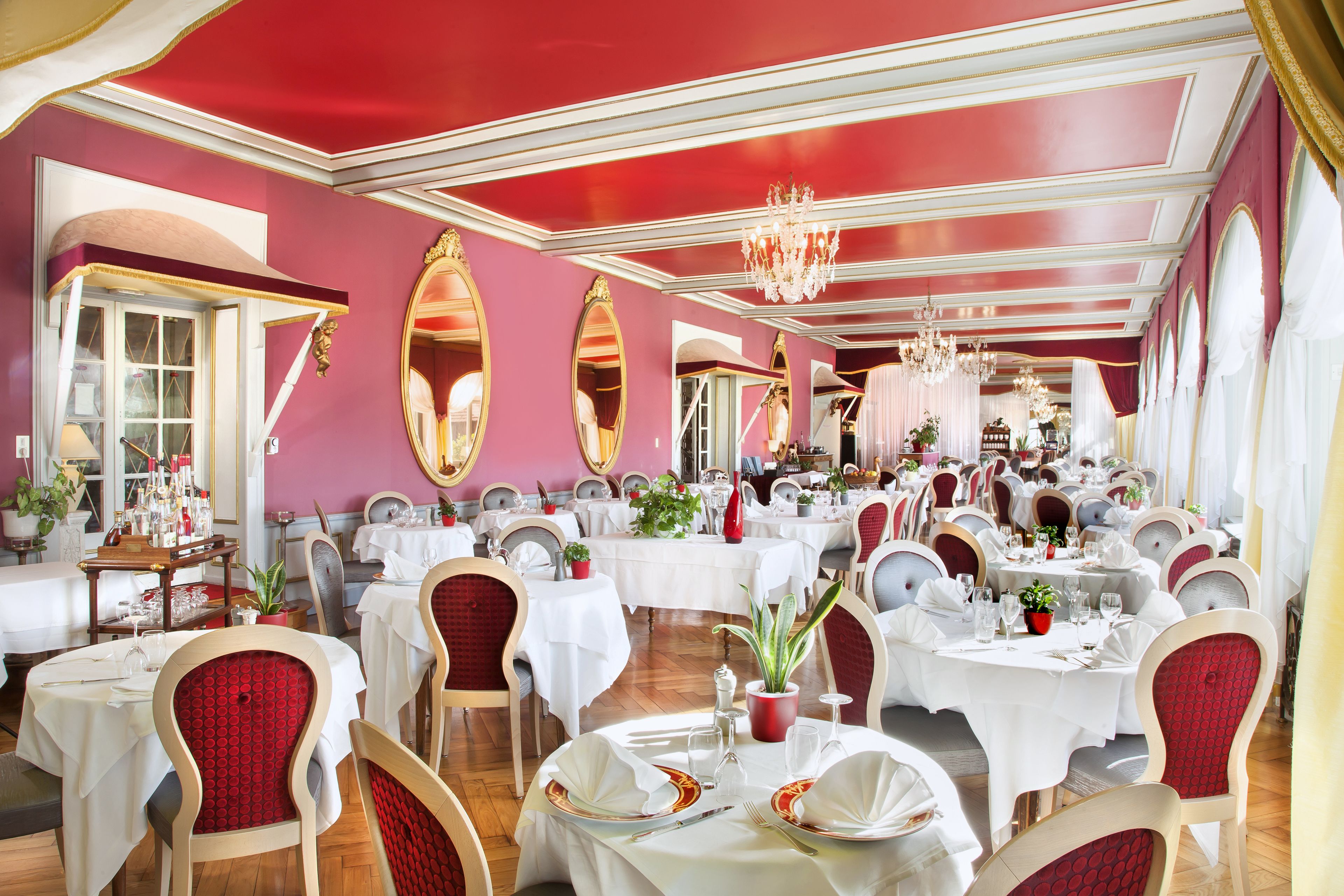 A spacious dining area with multiple round tables, each set with white tablecloths and red upholstered chairs.