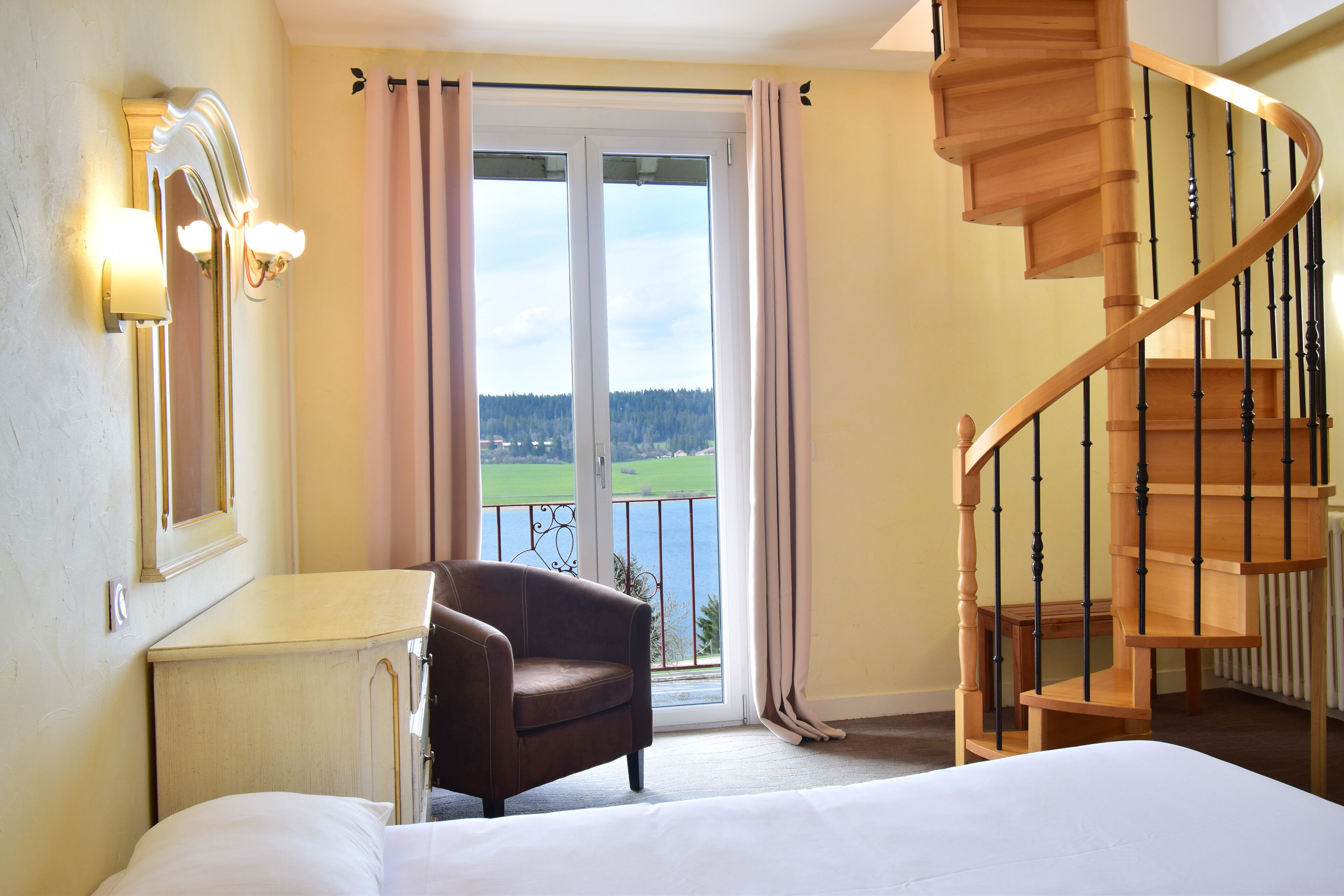 A bedroom with a bed, a chair, a desk, a mirror, a window with curtains, and a wooden staircase.