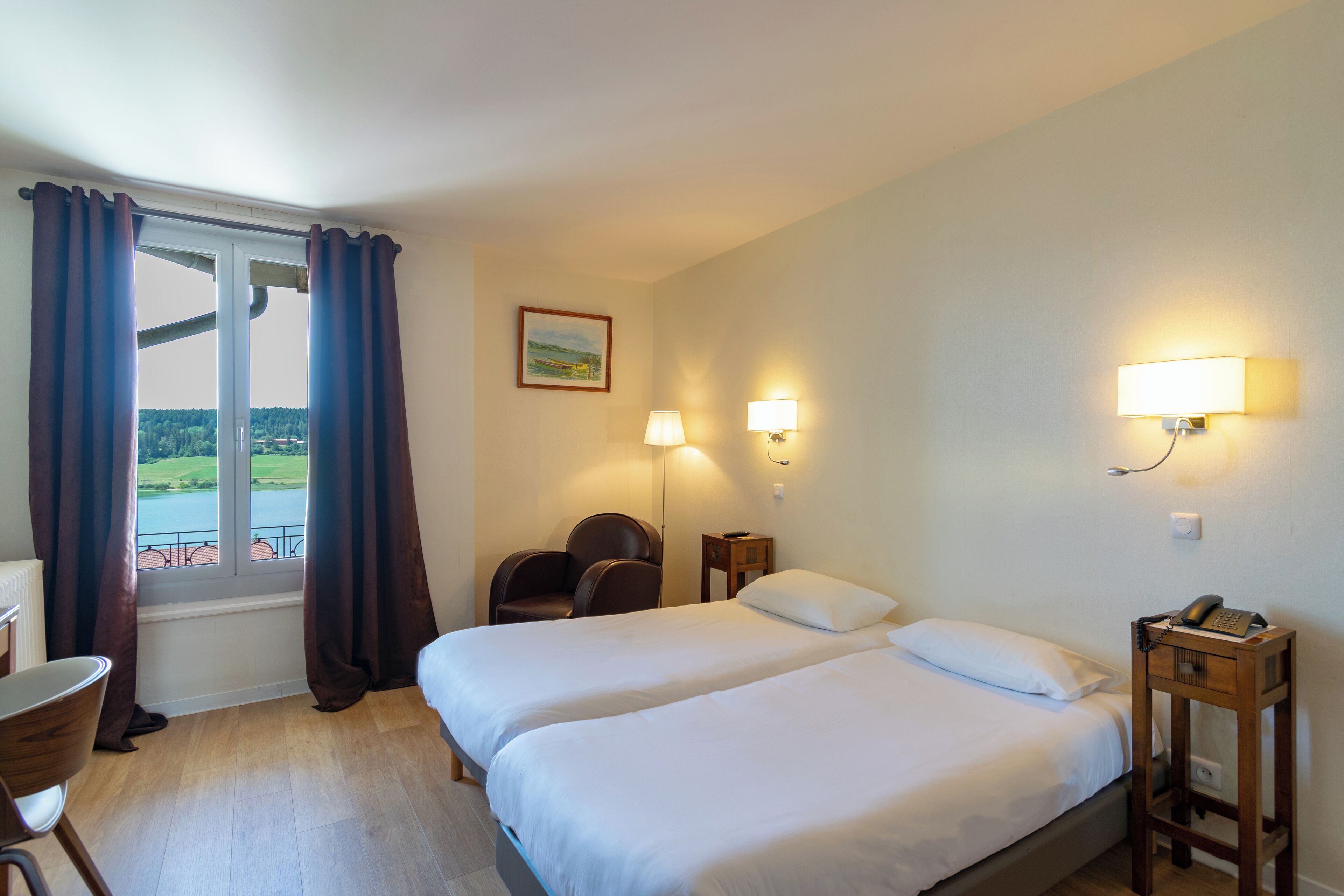 Hotelkamer met twee bedden, een bureau, een stoel, een lamp en uitzicht op een waterpartij.