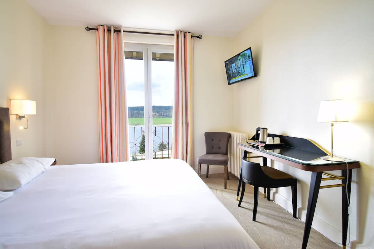 A hotel room with a large bed, a desk with a chair, a view of the outdoors through a window, and a painting on the wall.