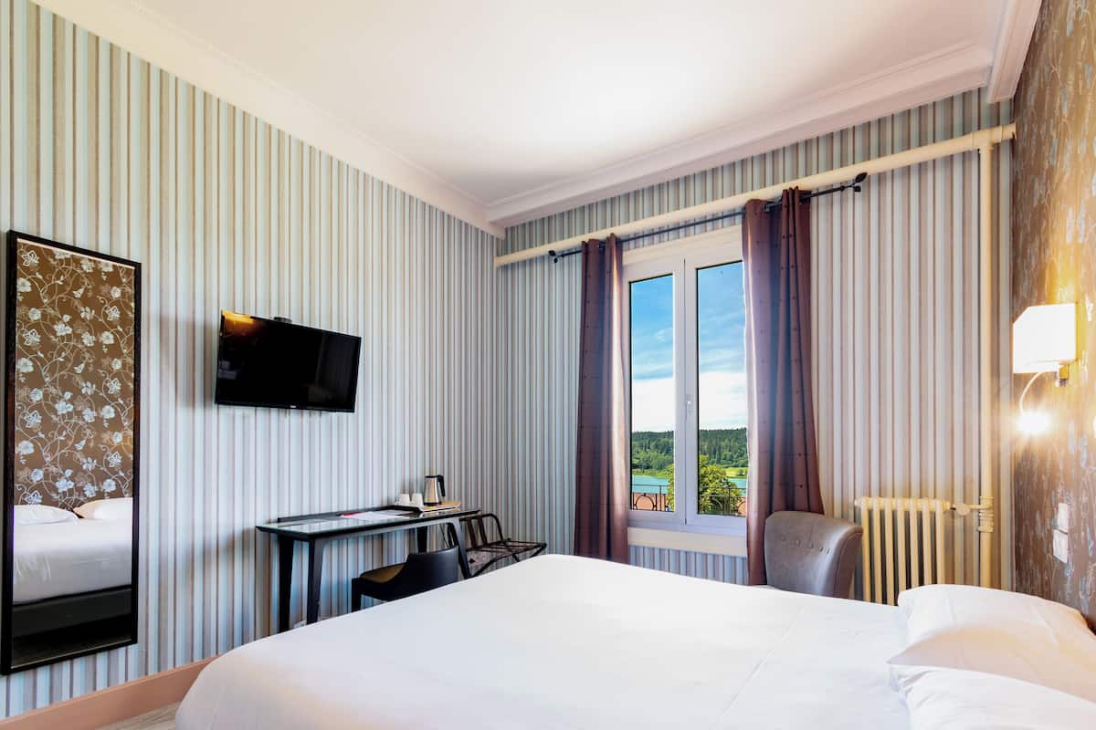 A hotel room with a bed, a desk with a chair, a TV, a window with curtains, and a radiator.