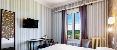 Habitación de hotel con cama, escritorio con silla, televisor, ventana con cortinas y radiador.
