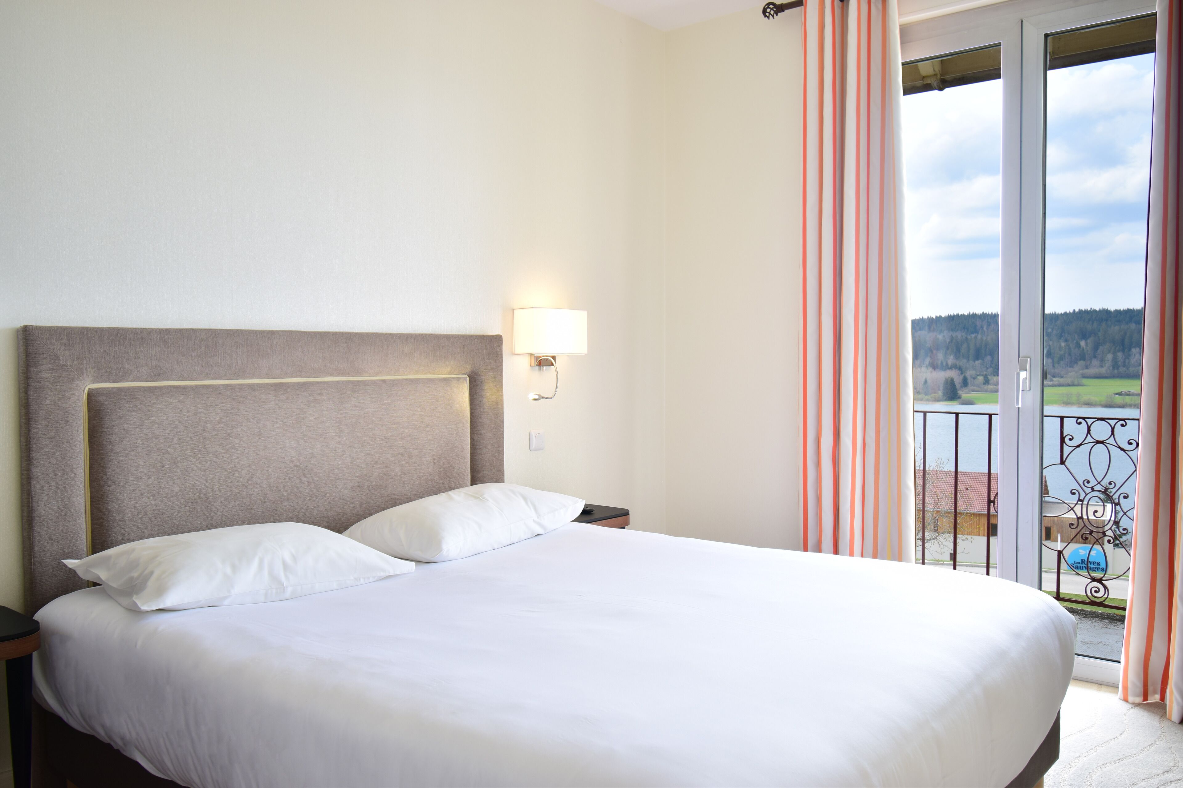 Une chambre d’hôtel avec un grand lit, une table de chevet, une lampe fixée au mur et un balcon offrant une vue sur la campagne.