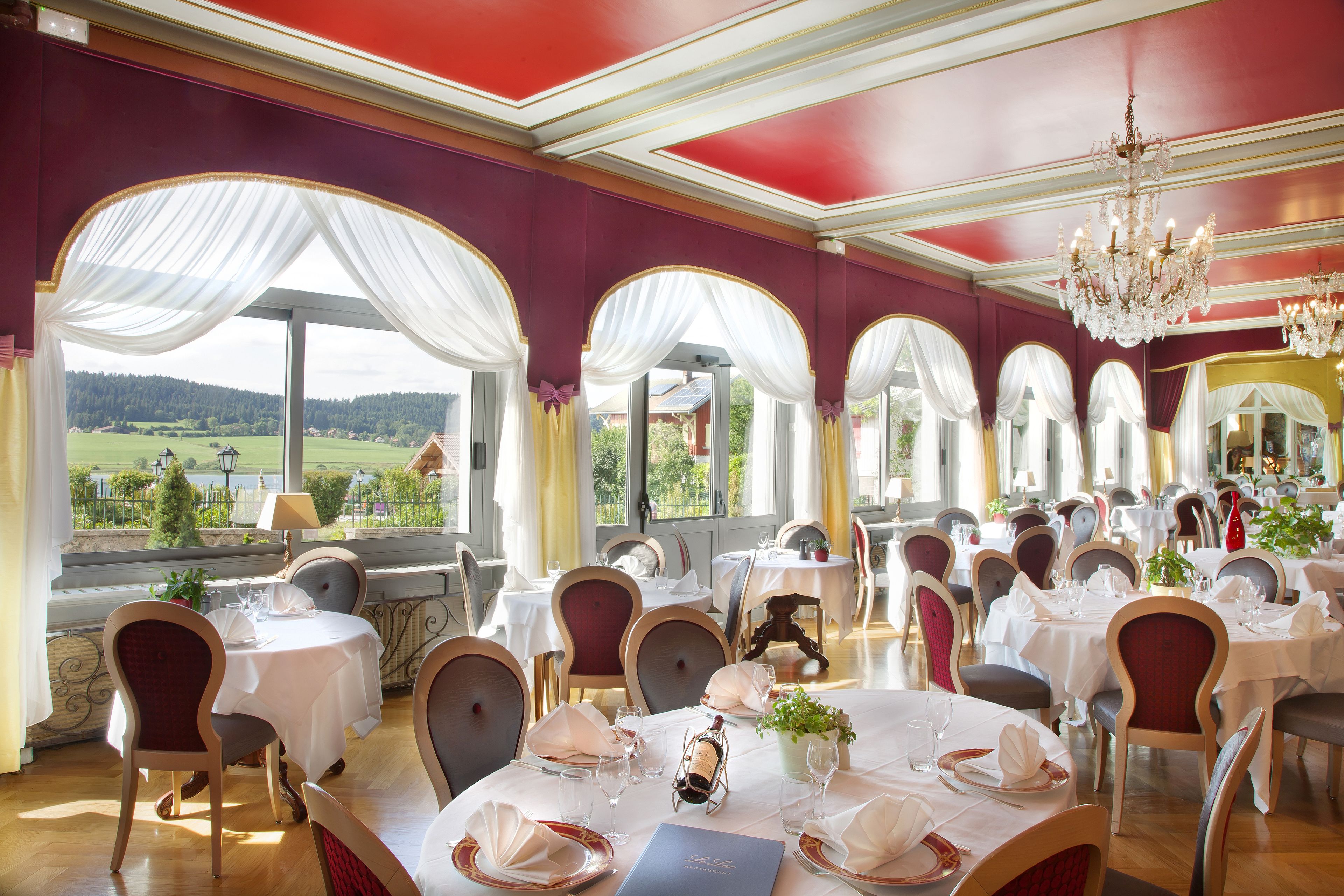 A dining area with tables set for service, large windows with draped curtains, and a chandelier.