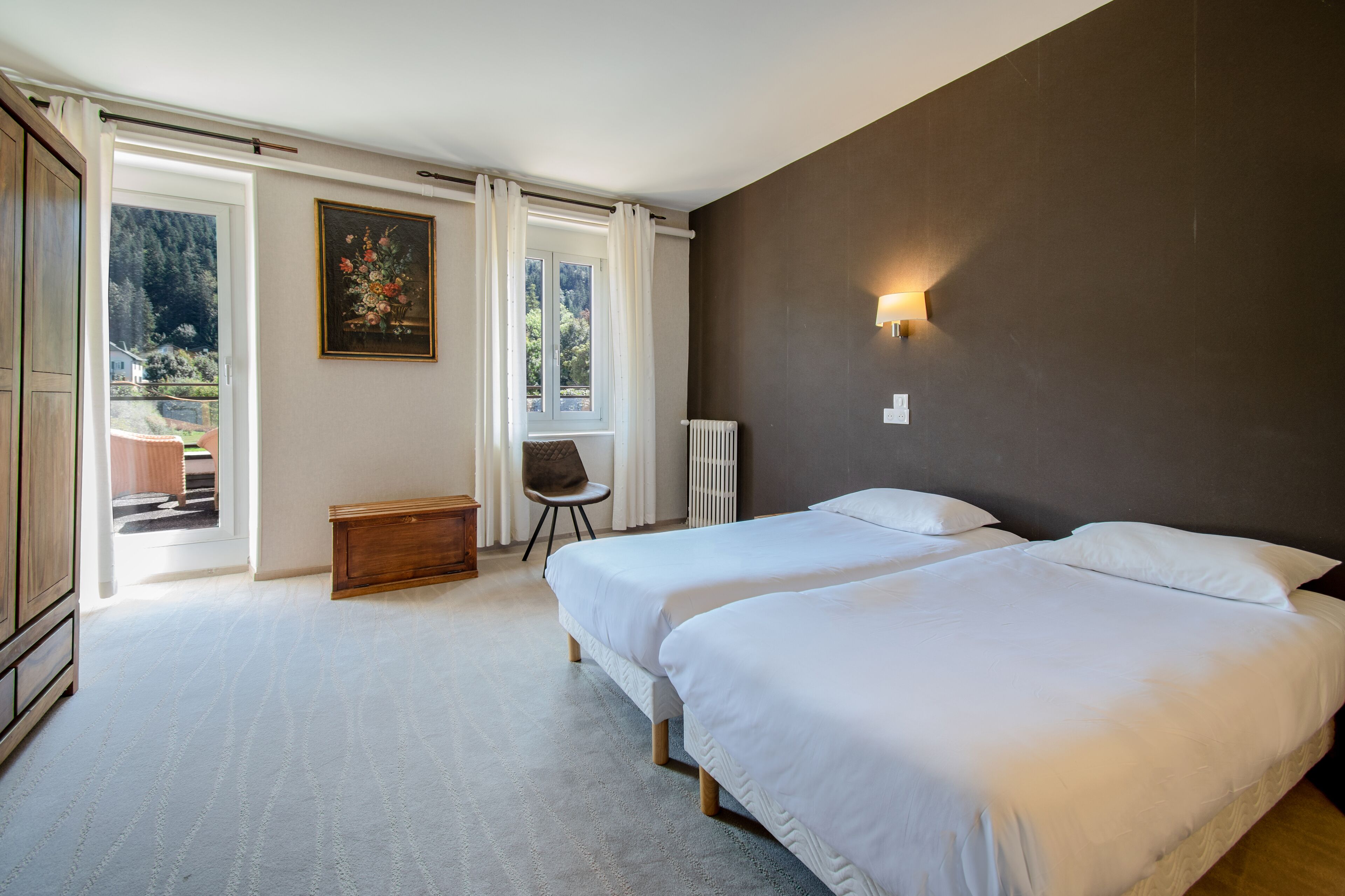 A bedroom with two beds, a wooden bench, a chair, a radiator, and a painting.